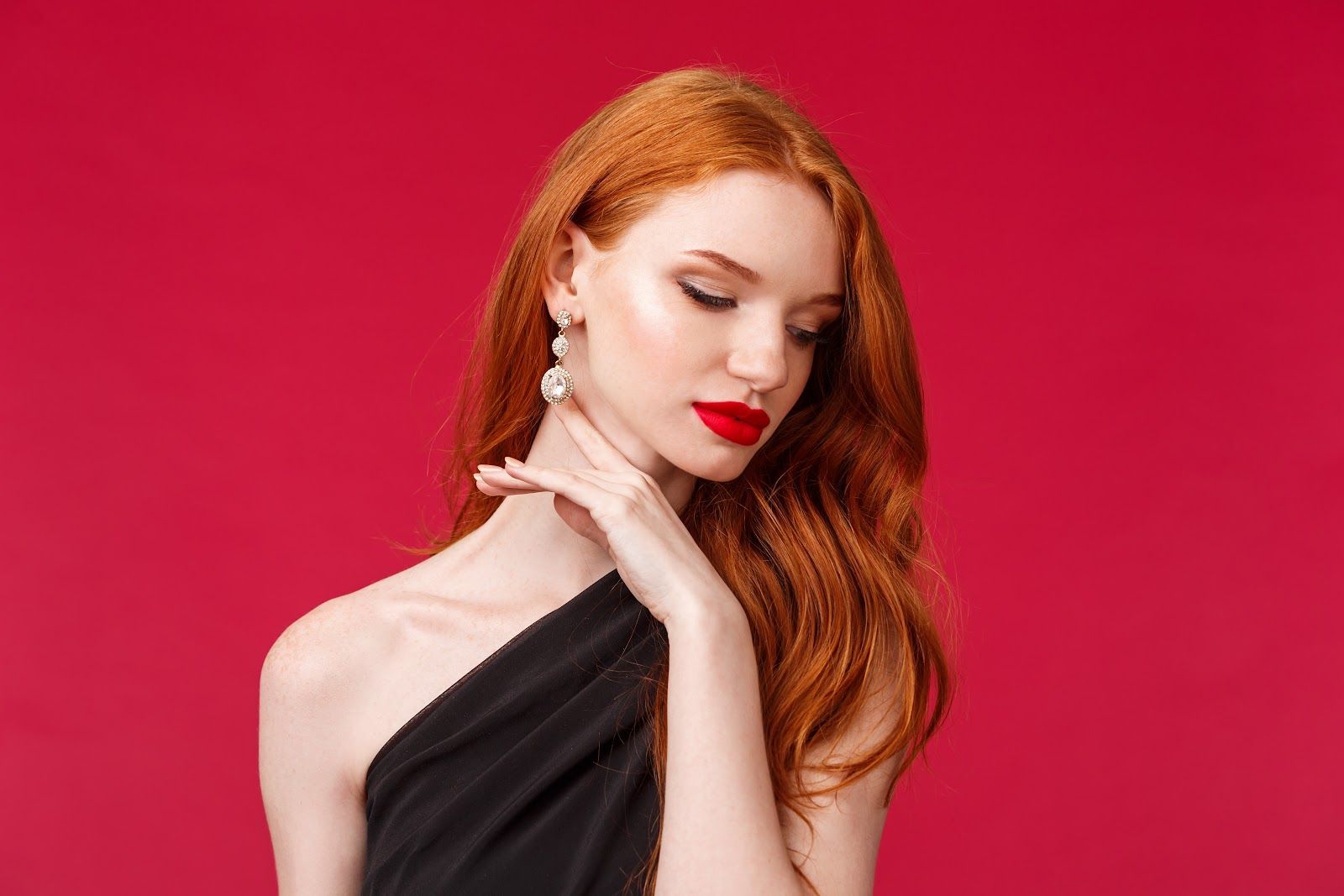 Woman with long red hair, wearing a black off-shoulder dress and pearl earrings, red lipstick, hand on neck, against red background.