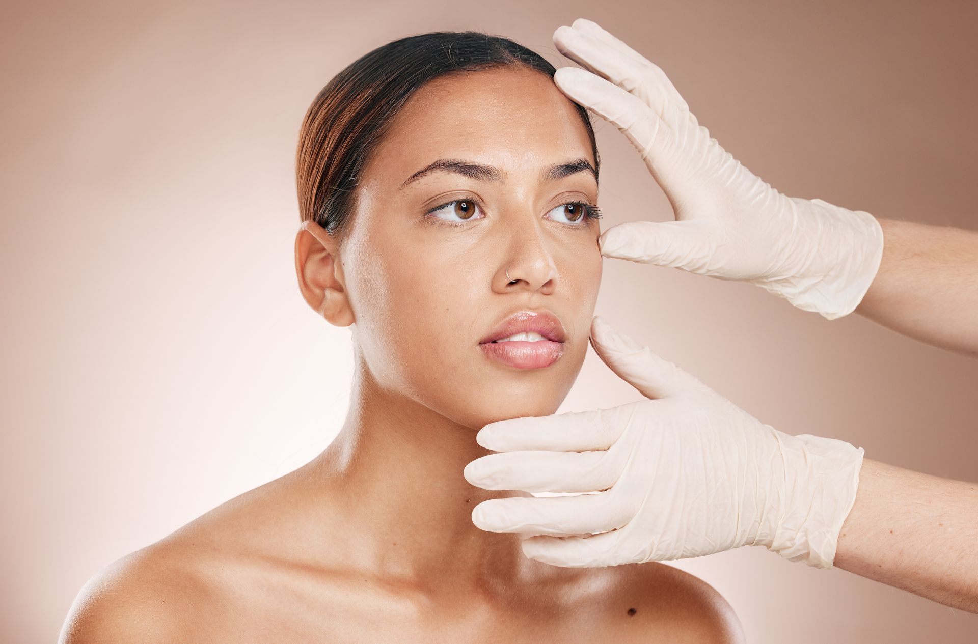 Woman's face examined by gloved hands, neutral expression, against a light brown background.