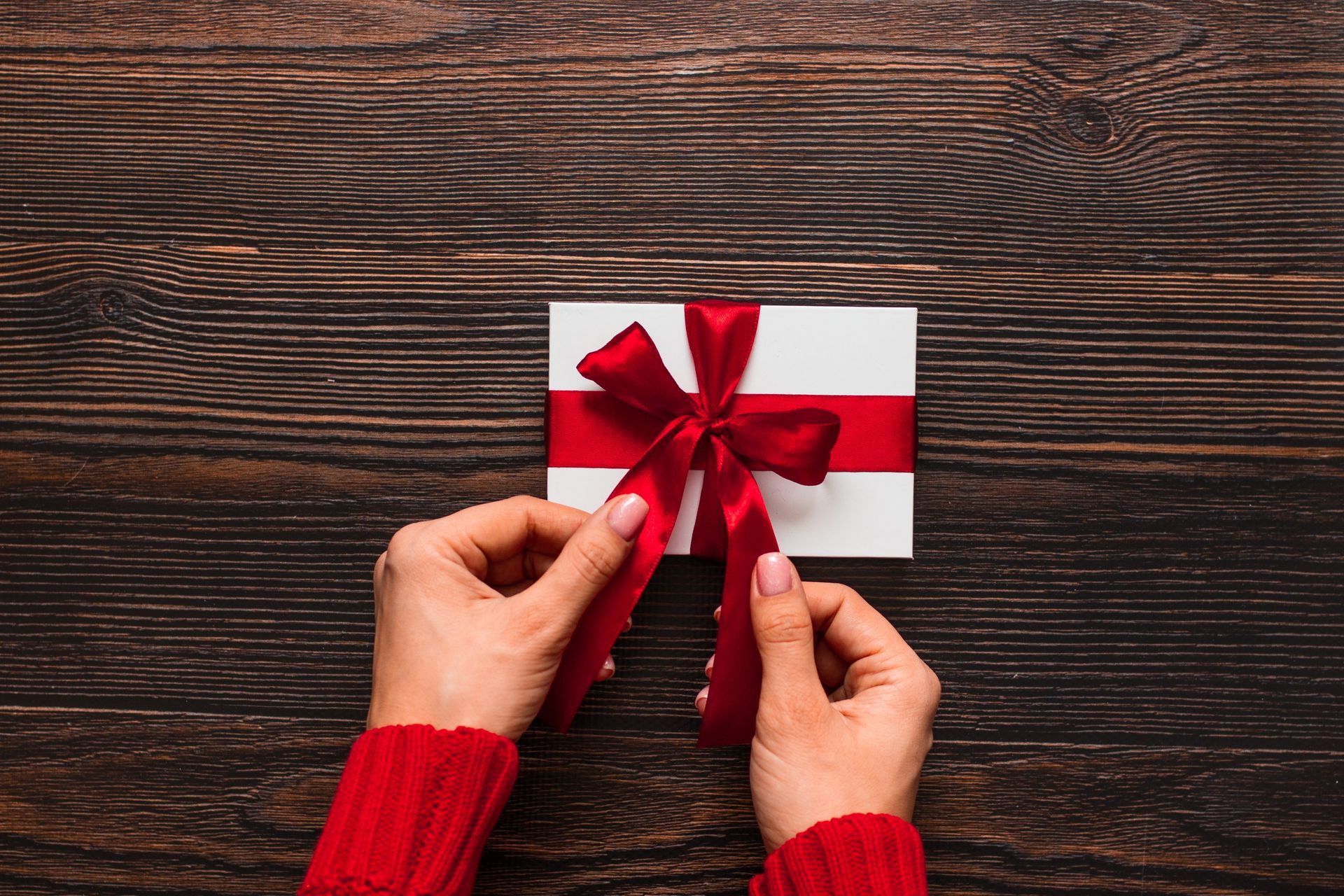 Hands tying a red ribbon on a white gift box against a dark wood background.
