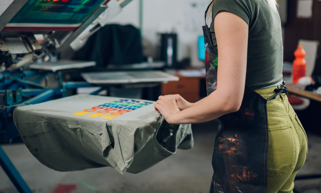 Person screen printing a colorful design on a grey t-shirt in a workshop, wearing a green shirt and apron.