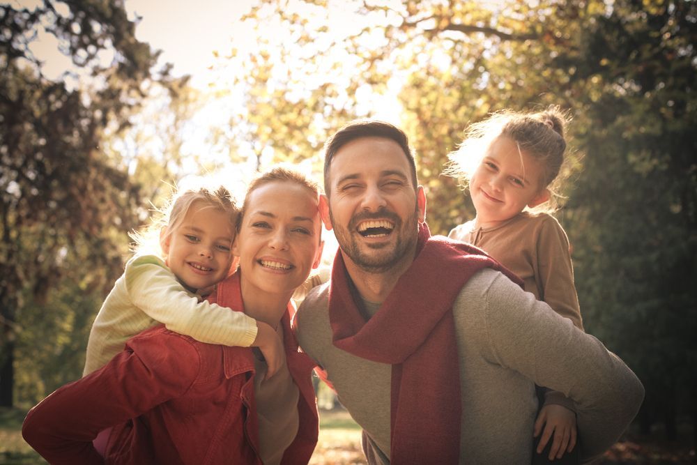 A smiling family outdoors in autumn, with parents giving their two children piggyback rides under warm sunlight.