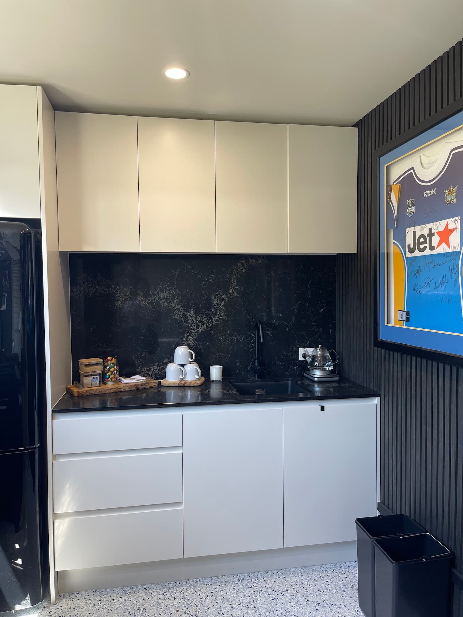Small modern kitchen with white cabinets, black countertop and backsplash, and framed artwork on dark textured wall.
