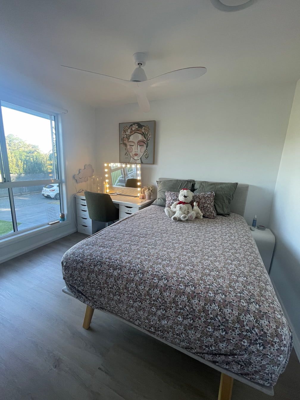 Bedroom with a bed, desk, and artwork. The bed has a floral patterned comforter. A large window lets in outside light.