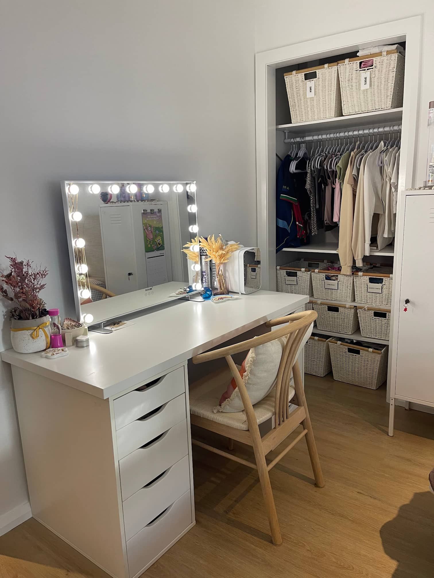 A white vanity desk with a lit mirror sits next to a closet filled with clothes and storage baskets.