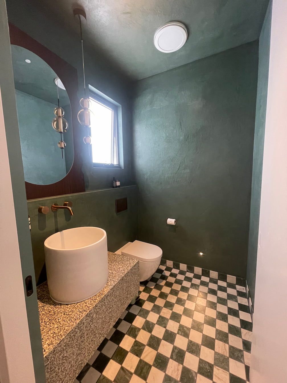 Small bathroom with green walls, checkered floor, round mirror, and a white sink.