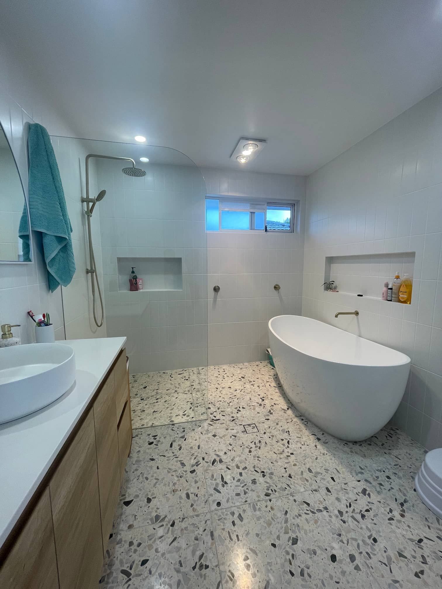 Modern white bathroom with oval bathtub and terrazzo tile floor. A wooden vanity with a round sink is on the left.