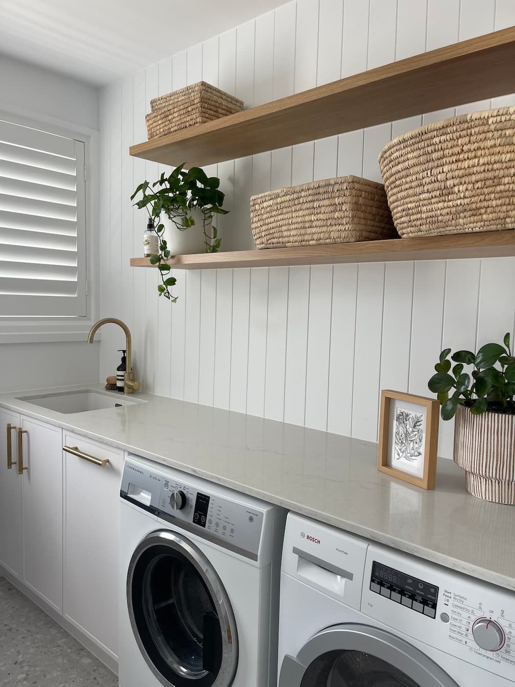 Laundry room with white cabinets, quartz countertop, washing machines, wooden shelves, and woven baskets.