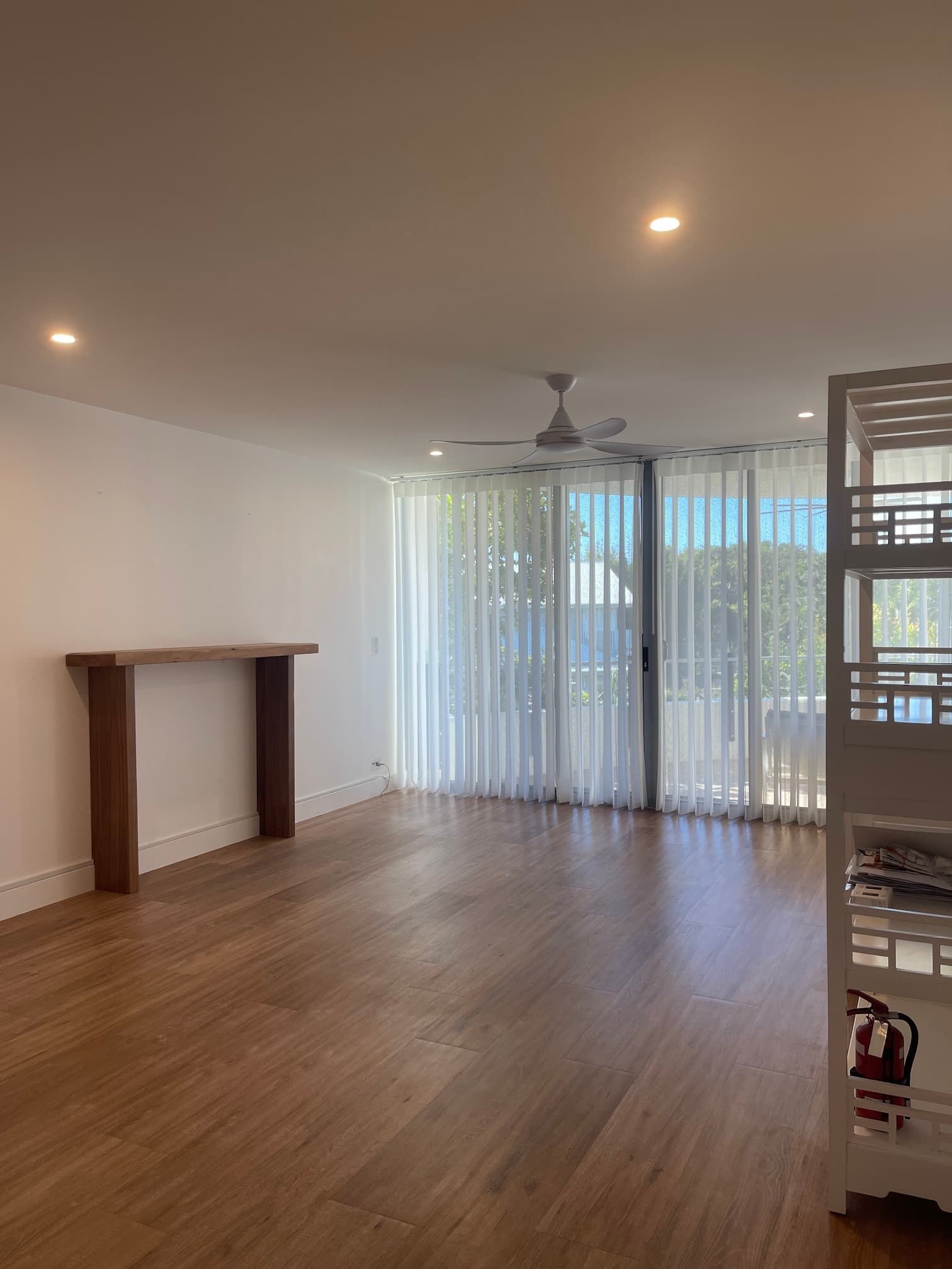 Empty room with wooden floor and a large window covered by vertical blinds, a wooden table against a white wall.