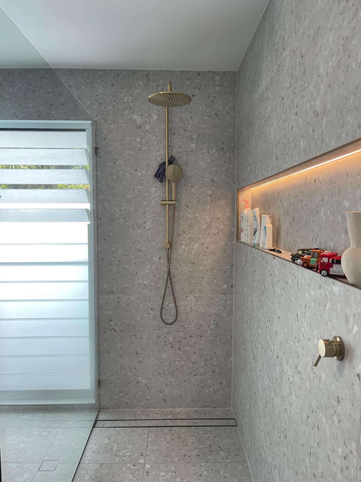 Gold shower fixture against gray speckled tile in a bathroom. Shelf with items lit by warm lighting.