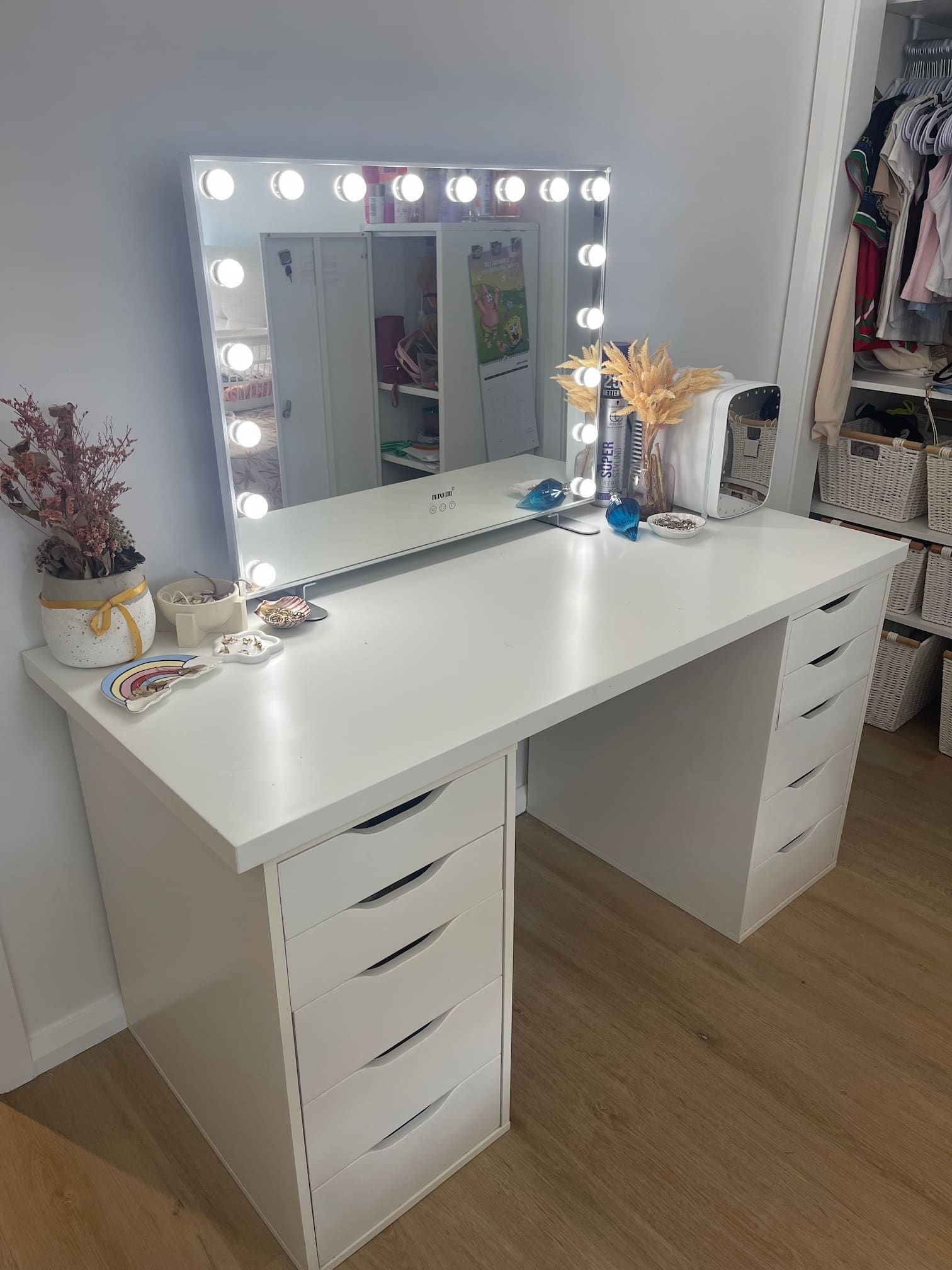 White vanity table with a mirror frame lit with lights, decorated with plants and makeup, in a room with a closet.