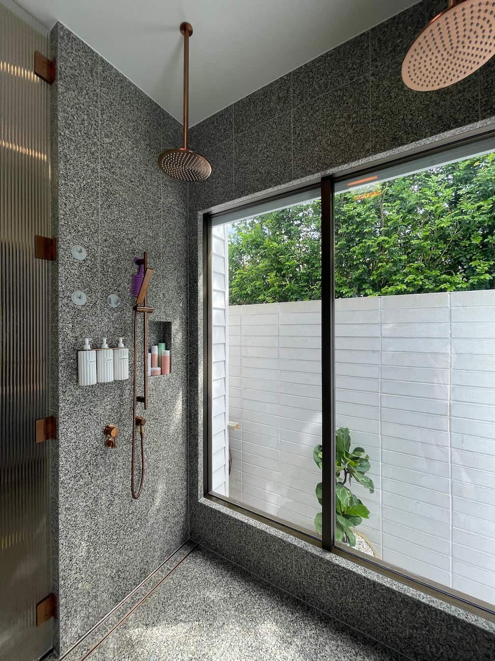 Shower with pebble tile, copper fixtures, large window, and foliage outside.