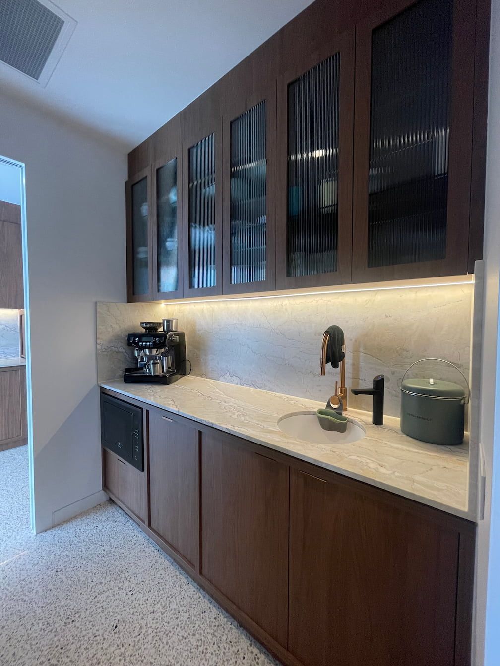 A dark wood butler's pantry with a coffee machine, sink, and cabinets with ribbed glass doors.