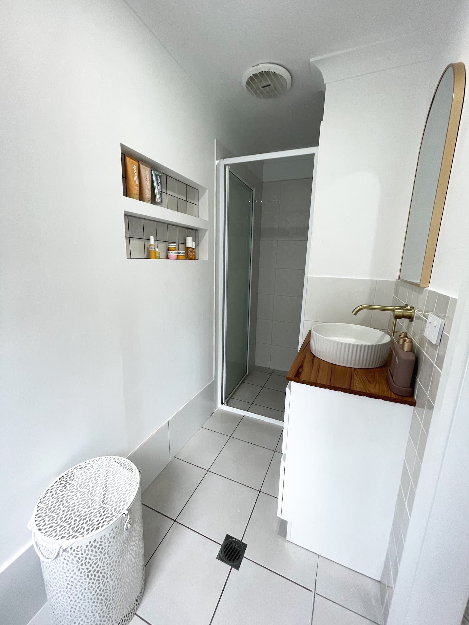 Small, white bathroom with a shower, sink, and built-in shelves. A white trash can sits near the entrance.