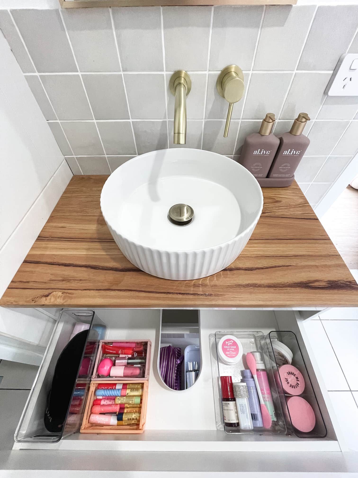 Bathroom sink with a wooden counter, gold fixtures, and a drawer filled with makeup organizers.