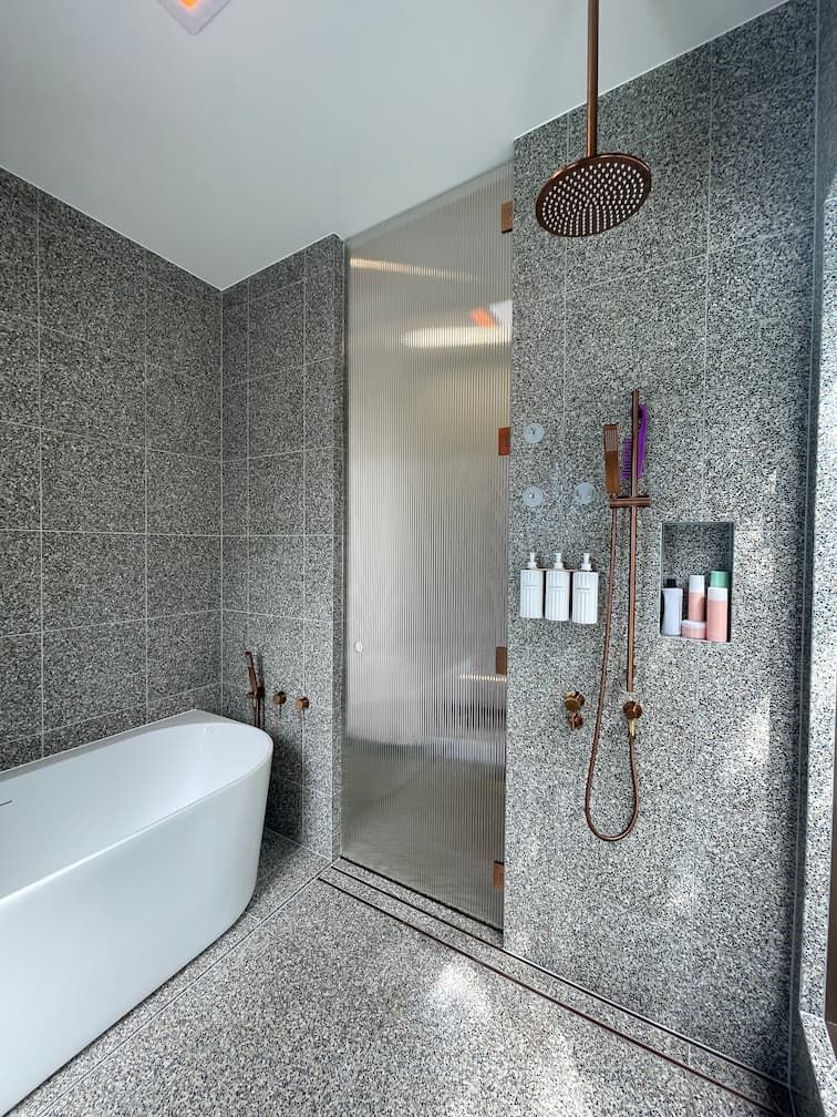 Bathroom with gray mosaic tile, a white tub, and a shower with copper fixtures.