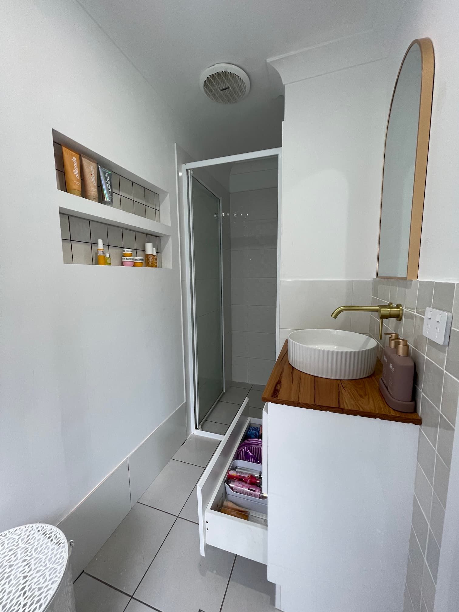 White bathroom with shower, sink, and storage; built-in niche with toiletries, open drawer with supplies.