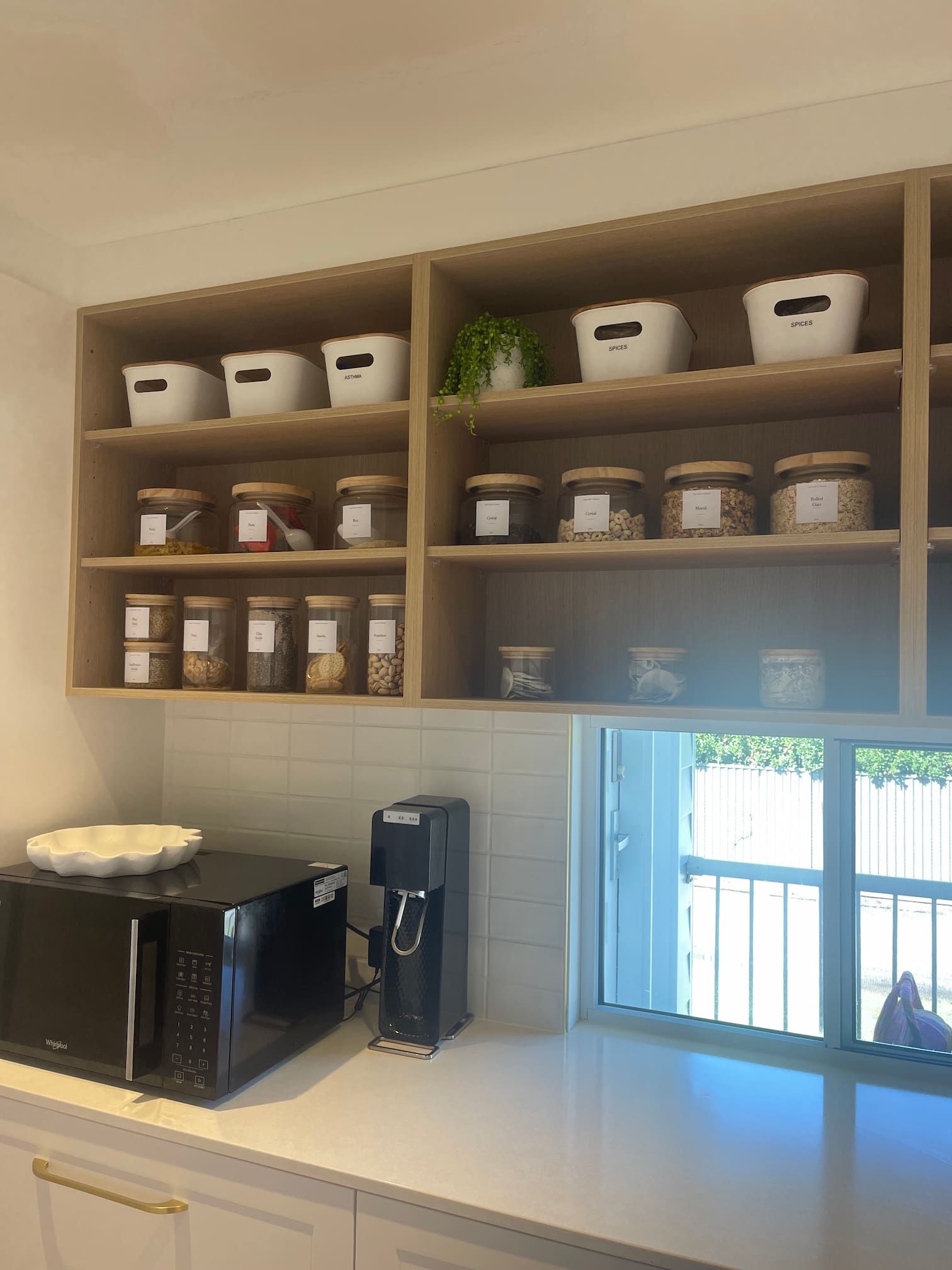 Kitchen shelving with jars of food and white containers. A microwave and water dispenser sit on the counter below a window.