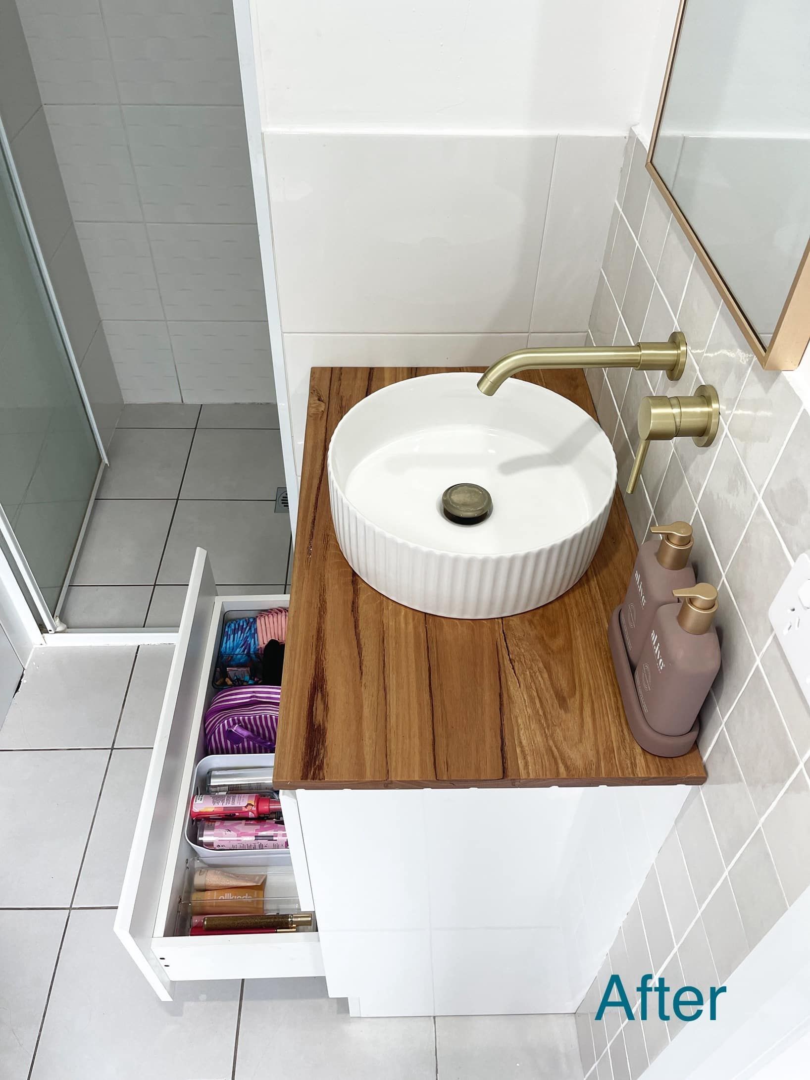White bathroom vanity with a wooden countertop and a round sink. Gold fixtures. Open drawer reveals folded clothes.