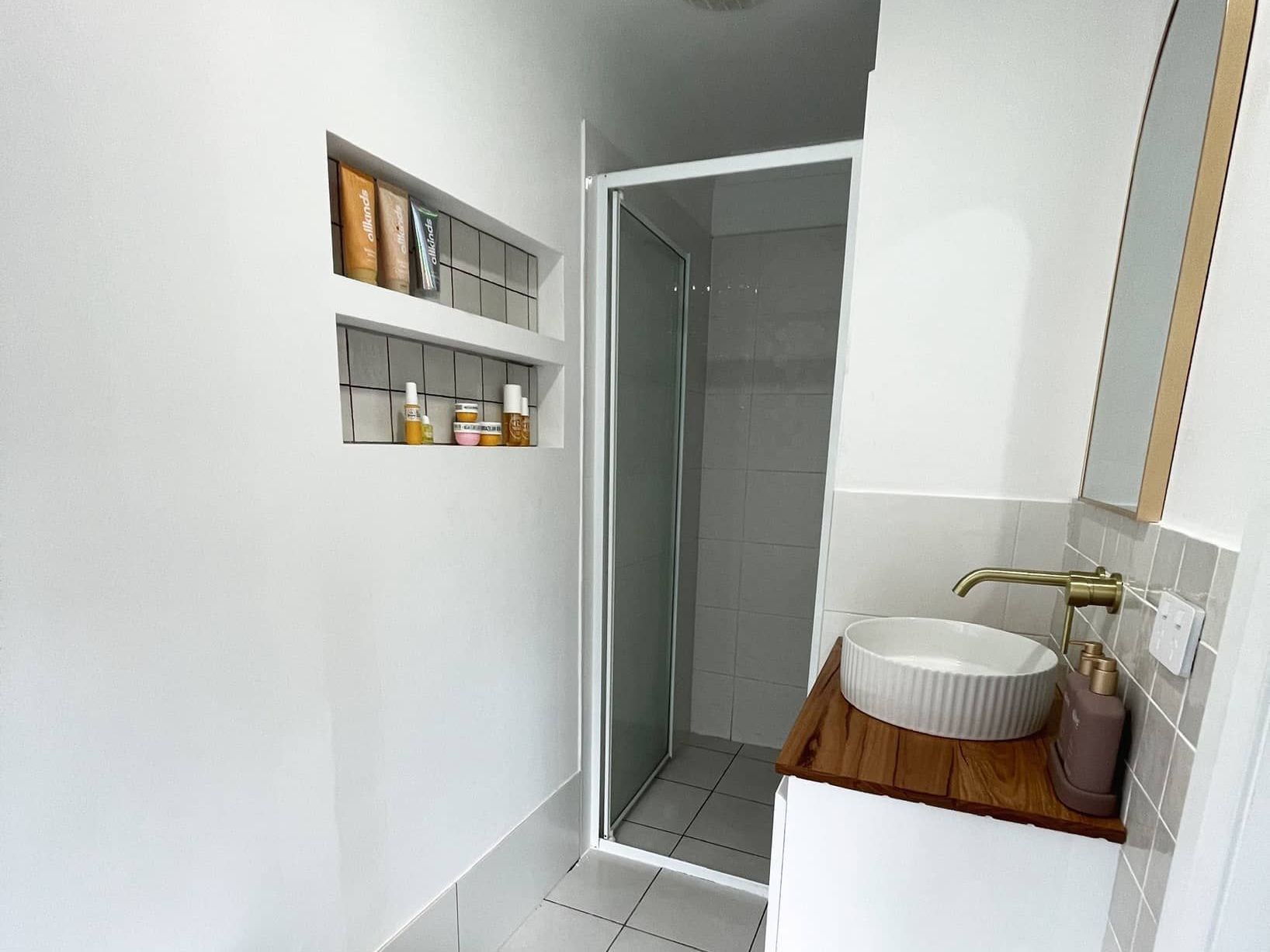 Small, modern bathroom with white walls, a shower, a vanity with a round sink, and a built-in shelf holding toiletries.