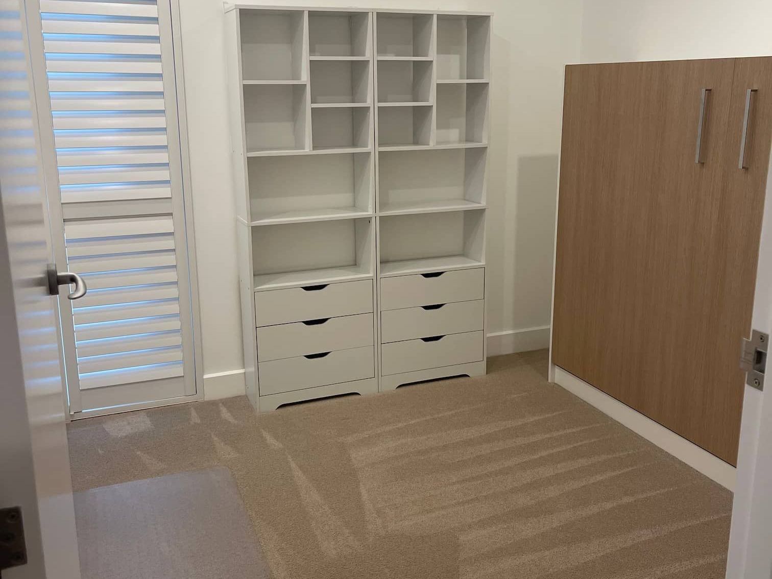 White shelving and drawers stand in a room with a shuttered door and wooden paneling.