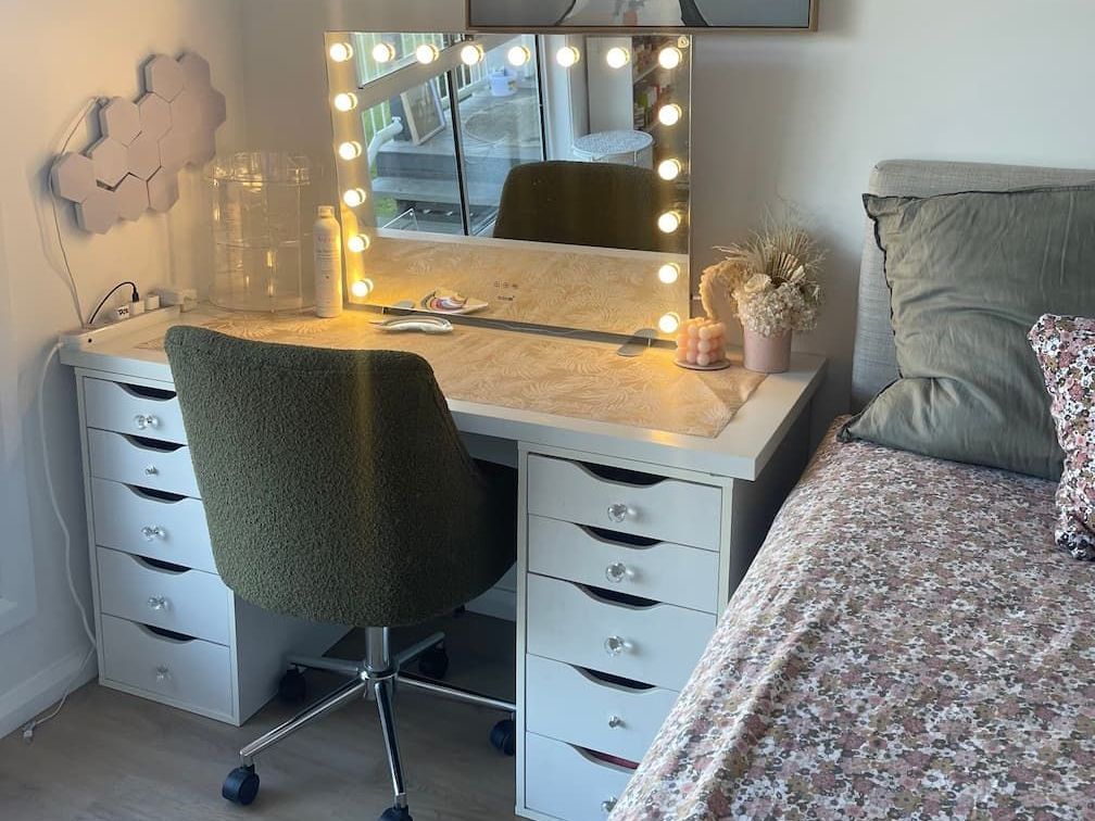 A white makeup vanity with a lighted mirror, green chair, and drawers, next to a bed with pillows.