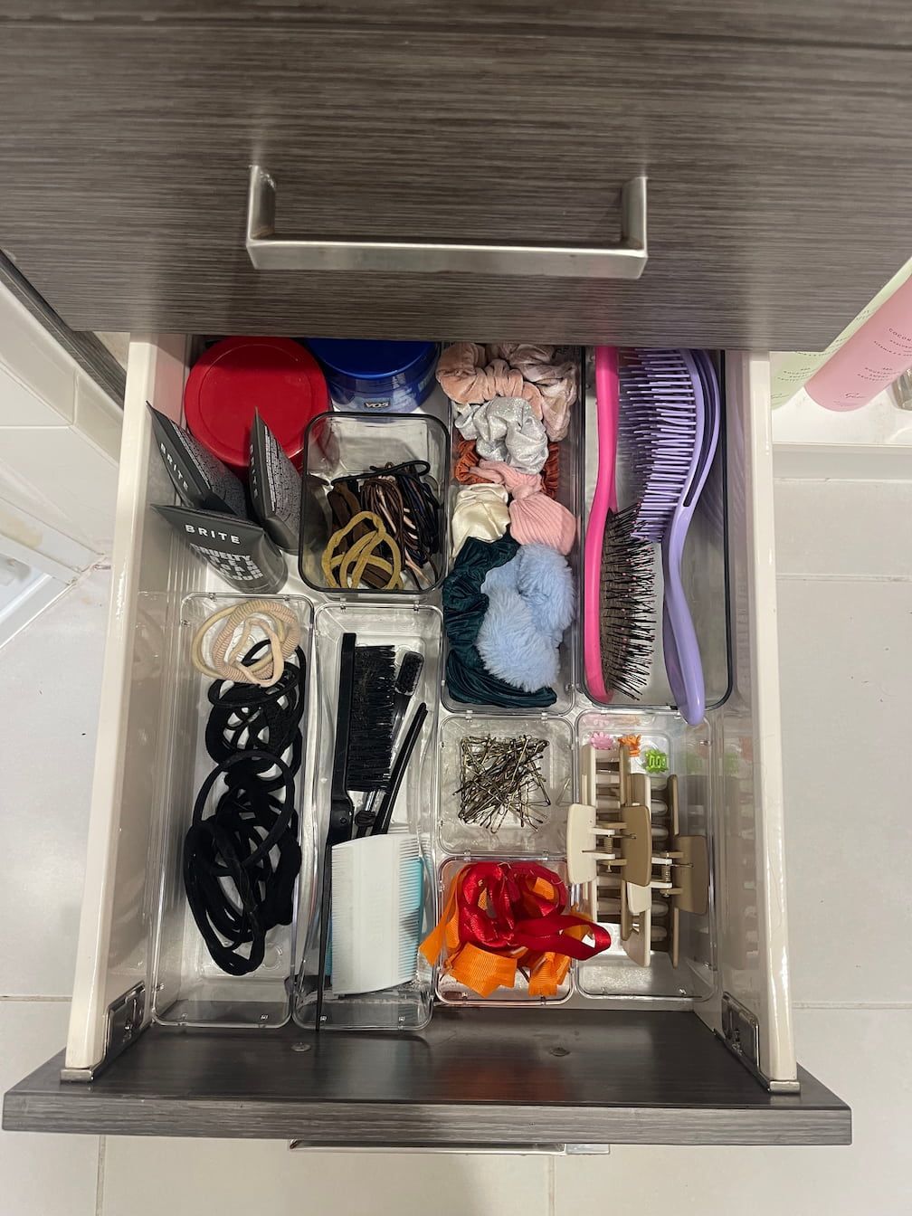 An open drawer with clear organizers filled with hair accessories, including elastics, clips, and brushes.