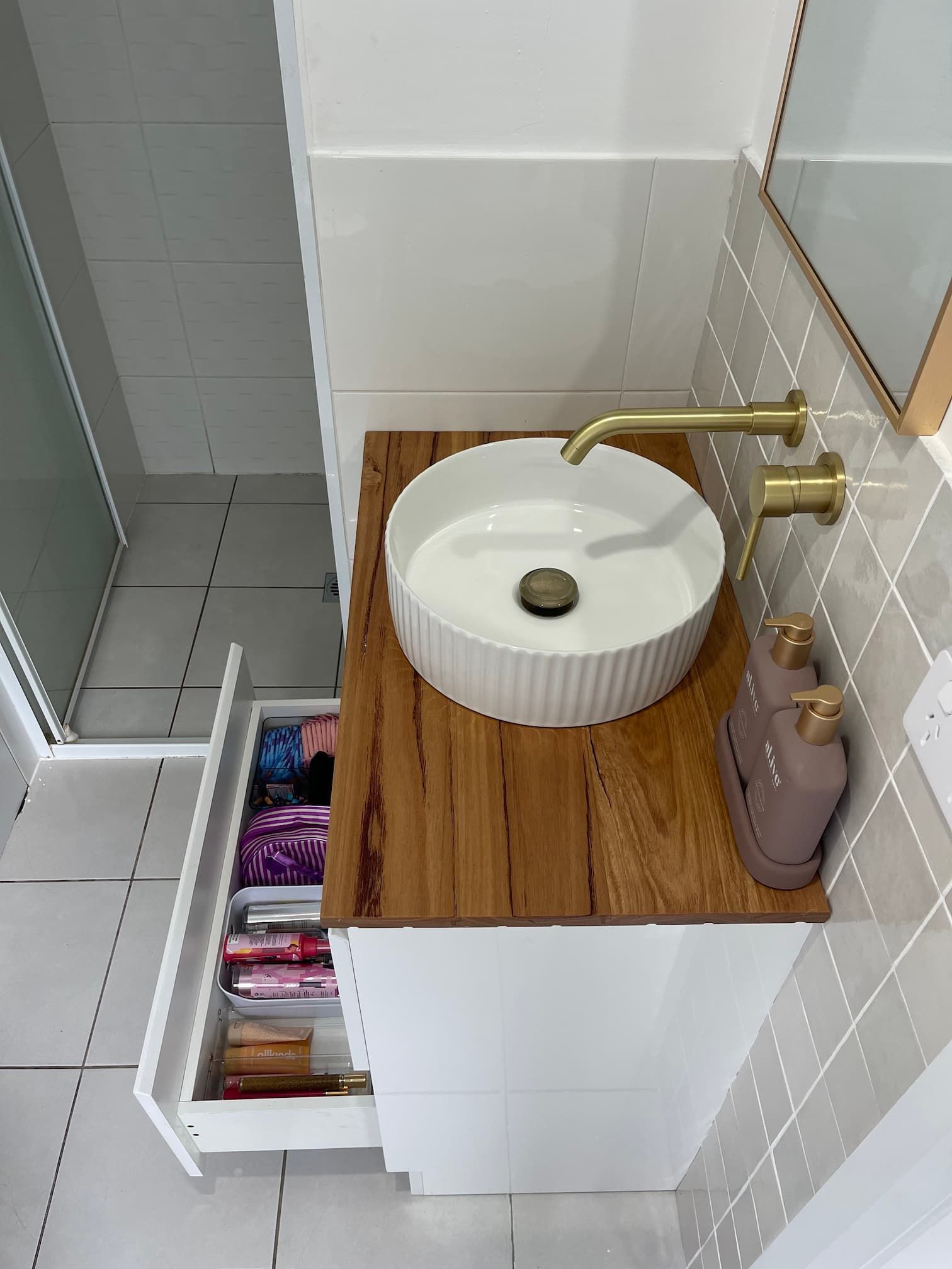 Bathroom vanity with white cabinet, wooden countertop, and round white sink. Brass faucet and matching soap dispensers.