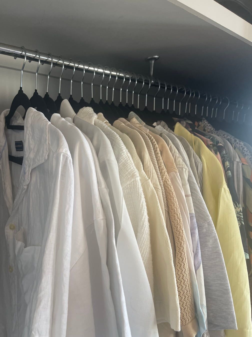 A closet with clothing hanging on a rod. Shirts range in color from white to yellow.