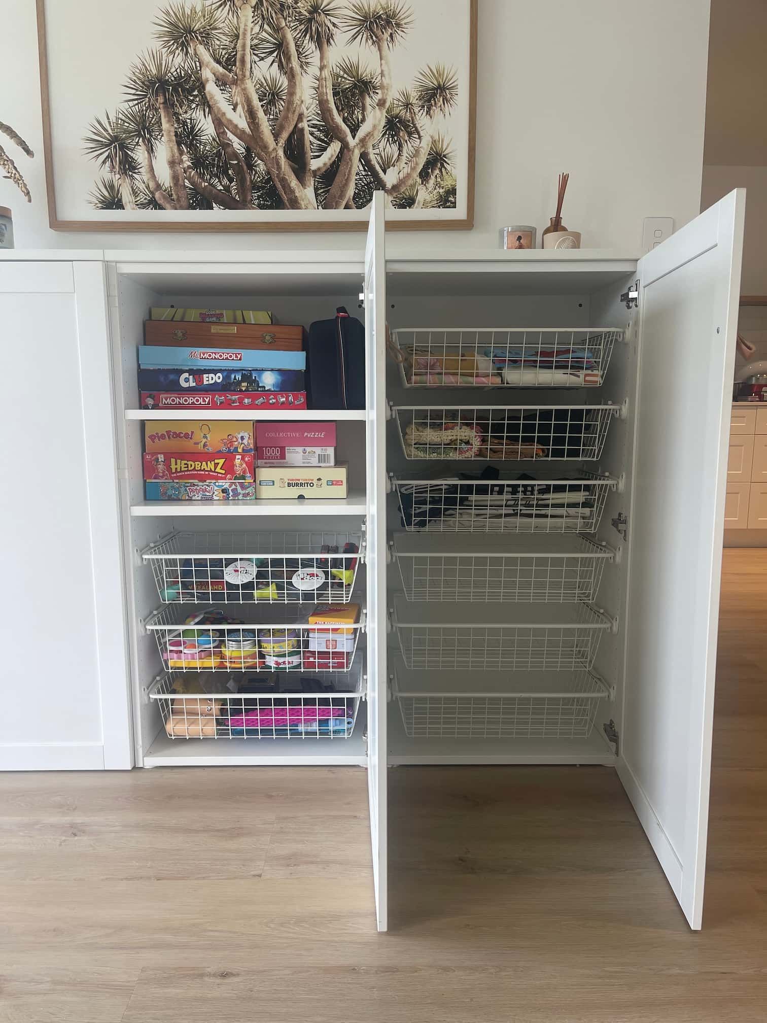 White cabinet with open door, showing shelves holding board games and wire baskets with colorful items.