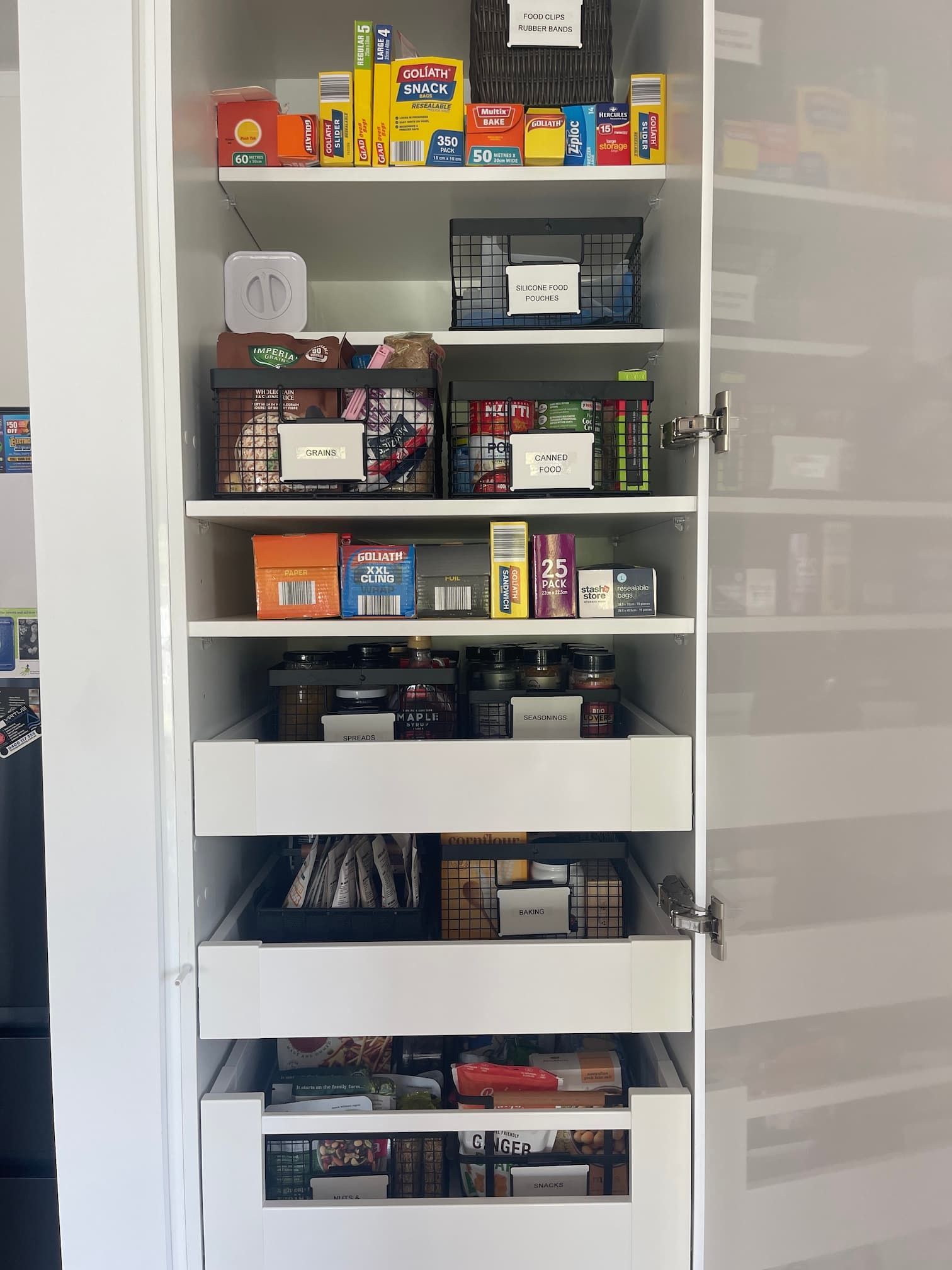 White pantry with organized shelves and pull-out drawers. Contains food items and other supplies in labeled containers.