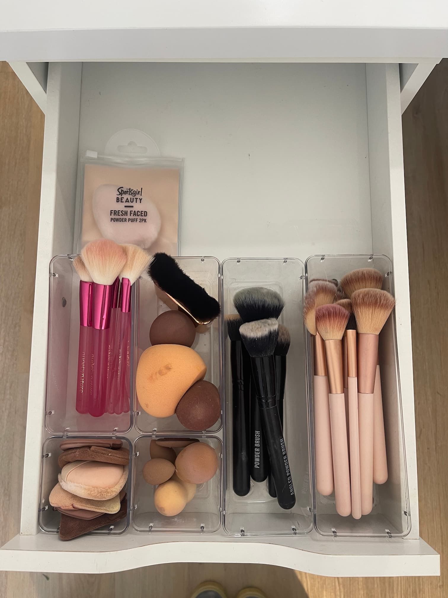 A drawer organized with clear plastic bins containing makeup brushes, sponges, and a powder puff.