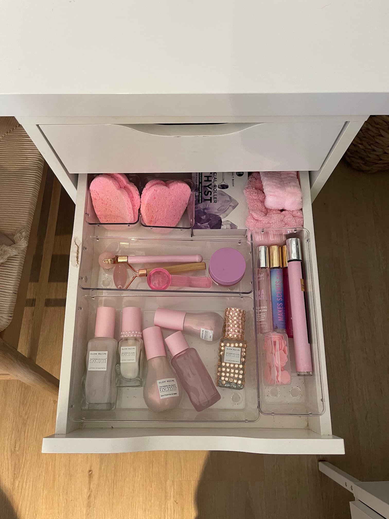 A white drawer filled with pink skincare and perfume bottles in clear organizers, along with pink heart-shaped sponges.