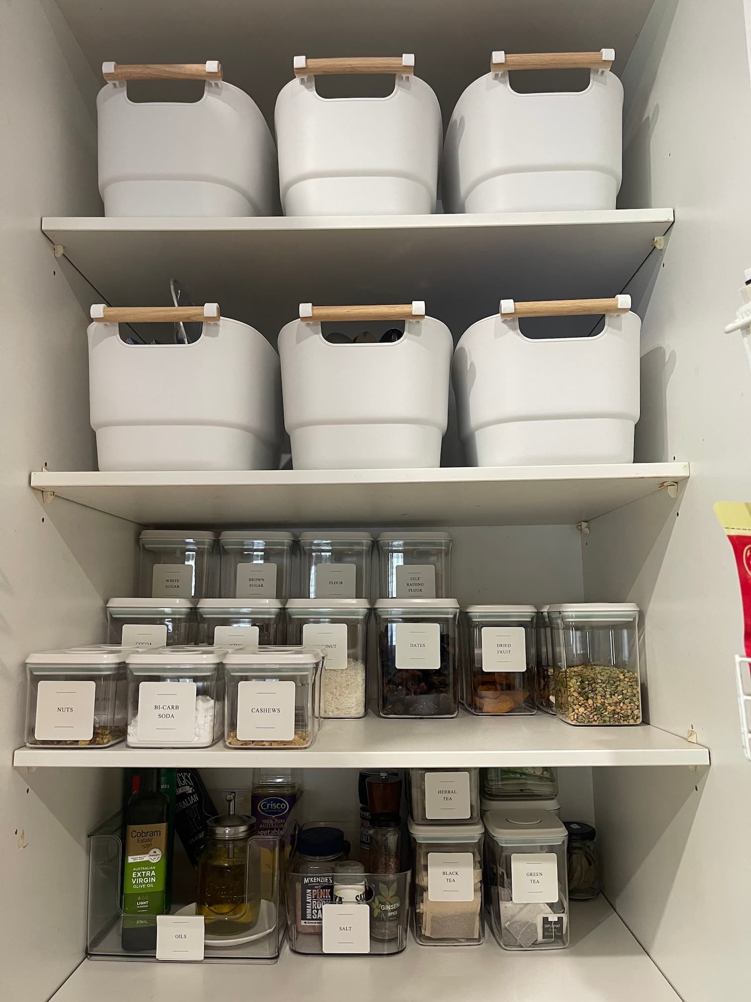 Organized pantry with white shelves. White storage bins with wooden handles are on the top shelves.