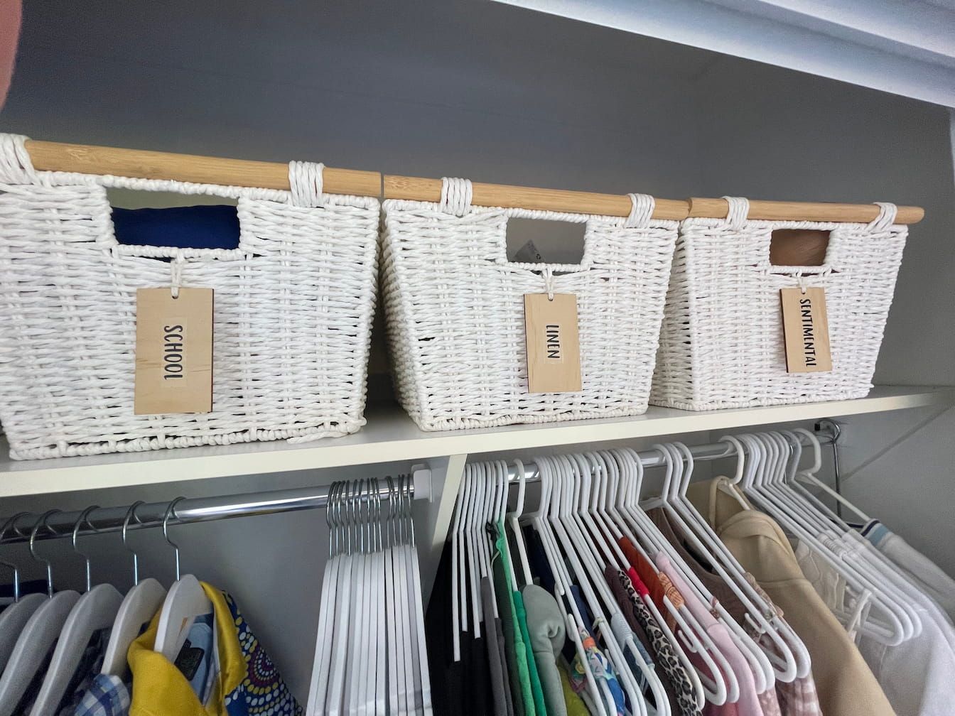 Three white wicker baskets with wooden handles on a shelf above a clothing rod.