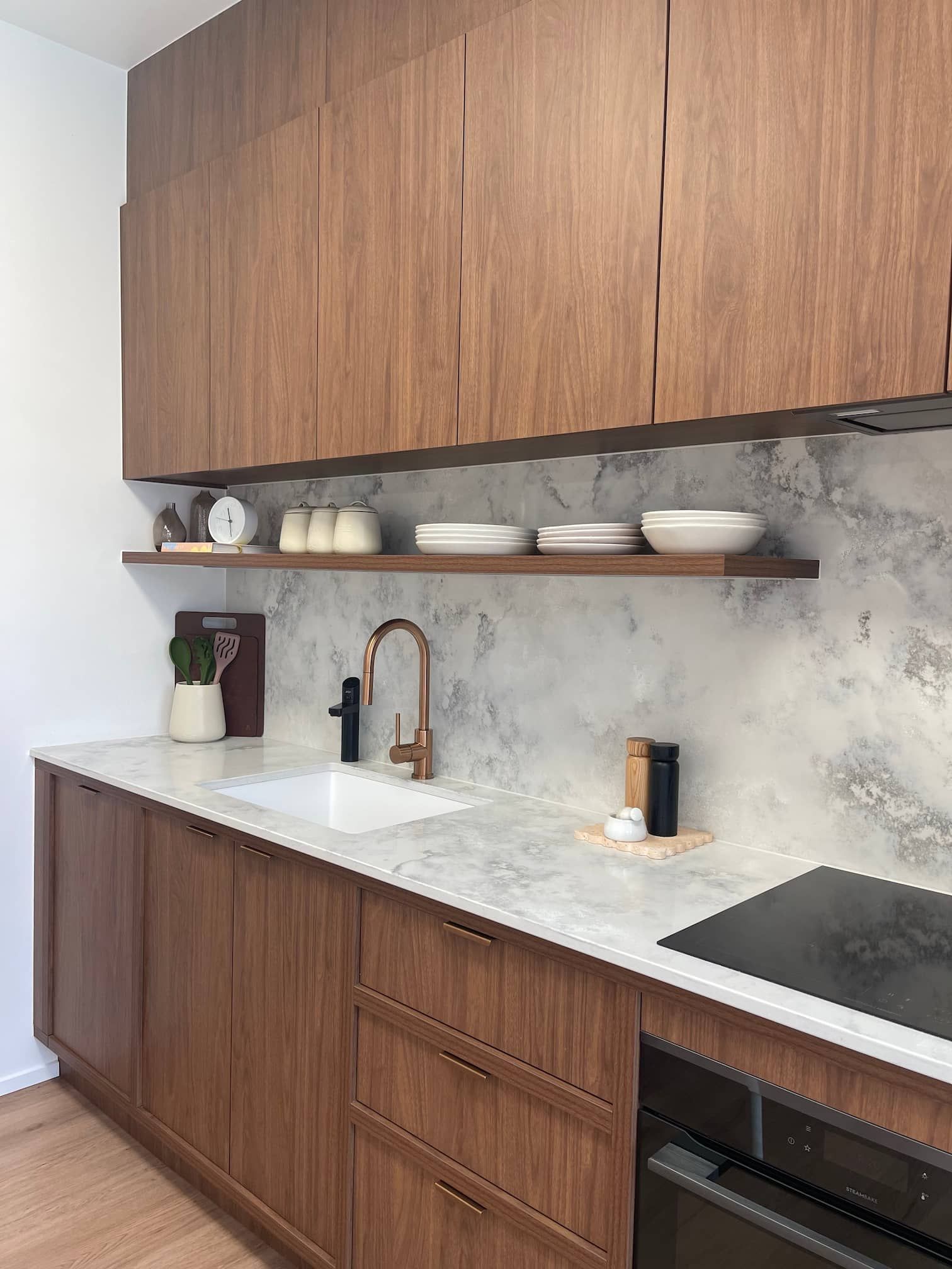 Modern kitchen with wood cabinets, marble backsplash, gold faucet, and floating shelf with dishes.