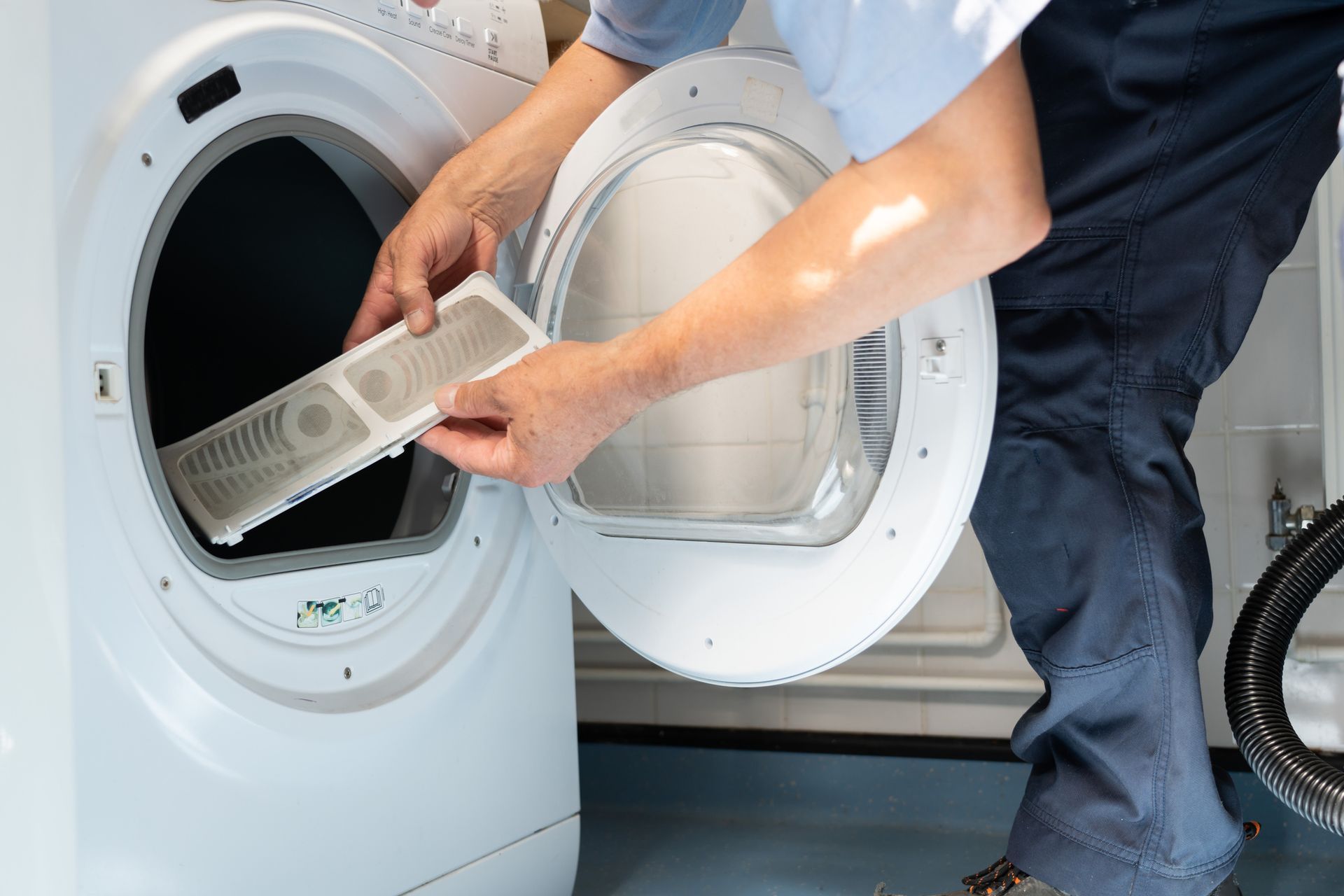 A person removes the lint filter from a white dryer, likely for cleaning. Close-up shot, well-lit room.