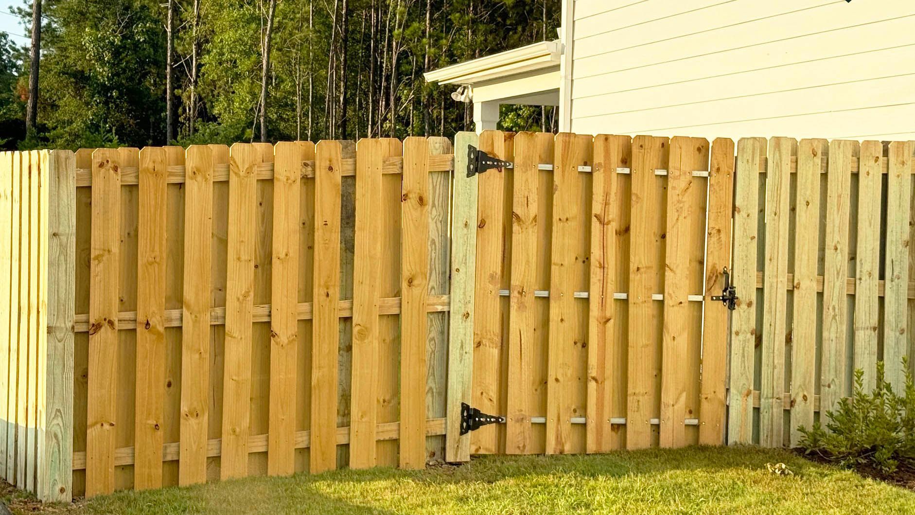 Wooden fence with a gate, outdoors. Brown wood, black hinges, green grass, and a house in the background.