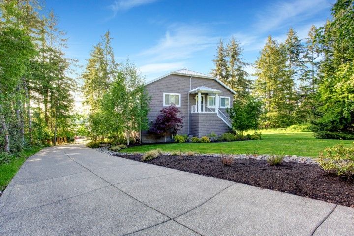 Gray house with a concrete driveway, surrounded by trees and green lawn under a blue sky.