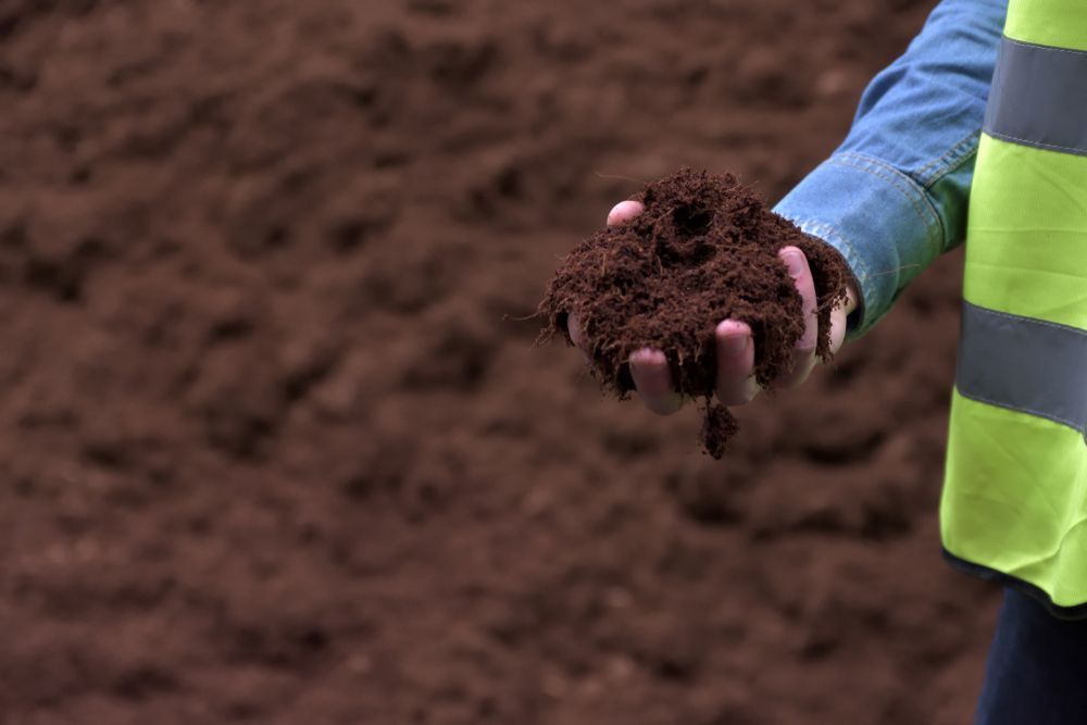 Person in safety vest holds a handful of dark brown, loamy soil. A large pile of soil is in the background.