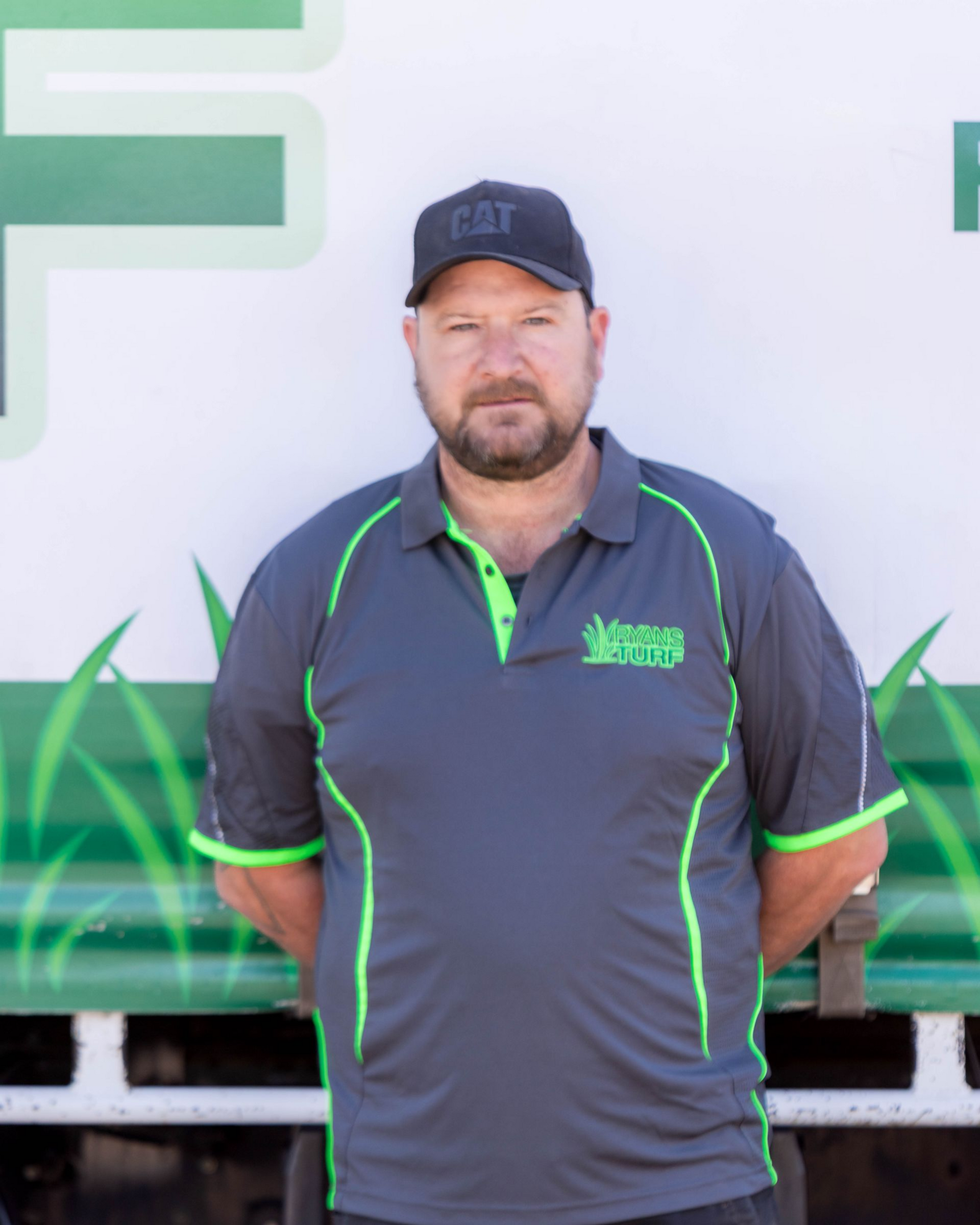 A man wearing a black CAT cap and a gray polo shirt with neon green trim stands against a backdrop with green grass art.