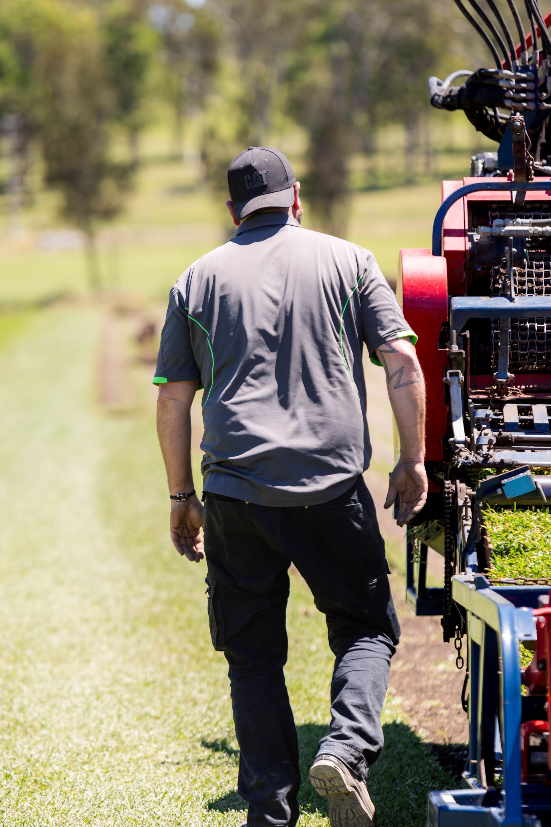Man in gray shirt and black pants walks beside a machine laying sod on green grass.
