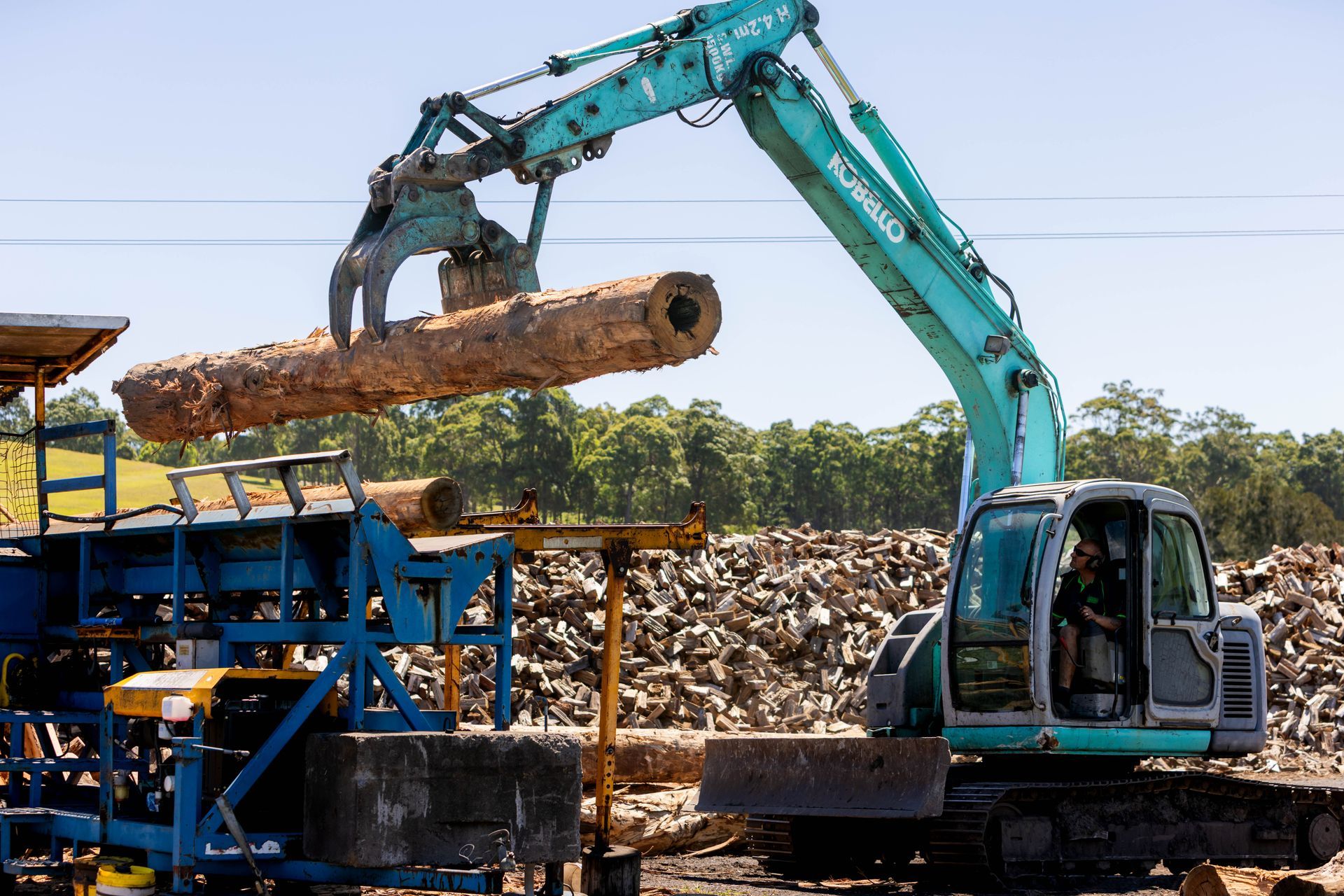 Excavator lifting a log, placing it on a conveyor belt at a lumber mill.