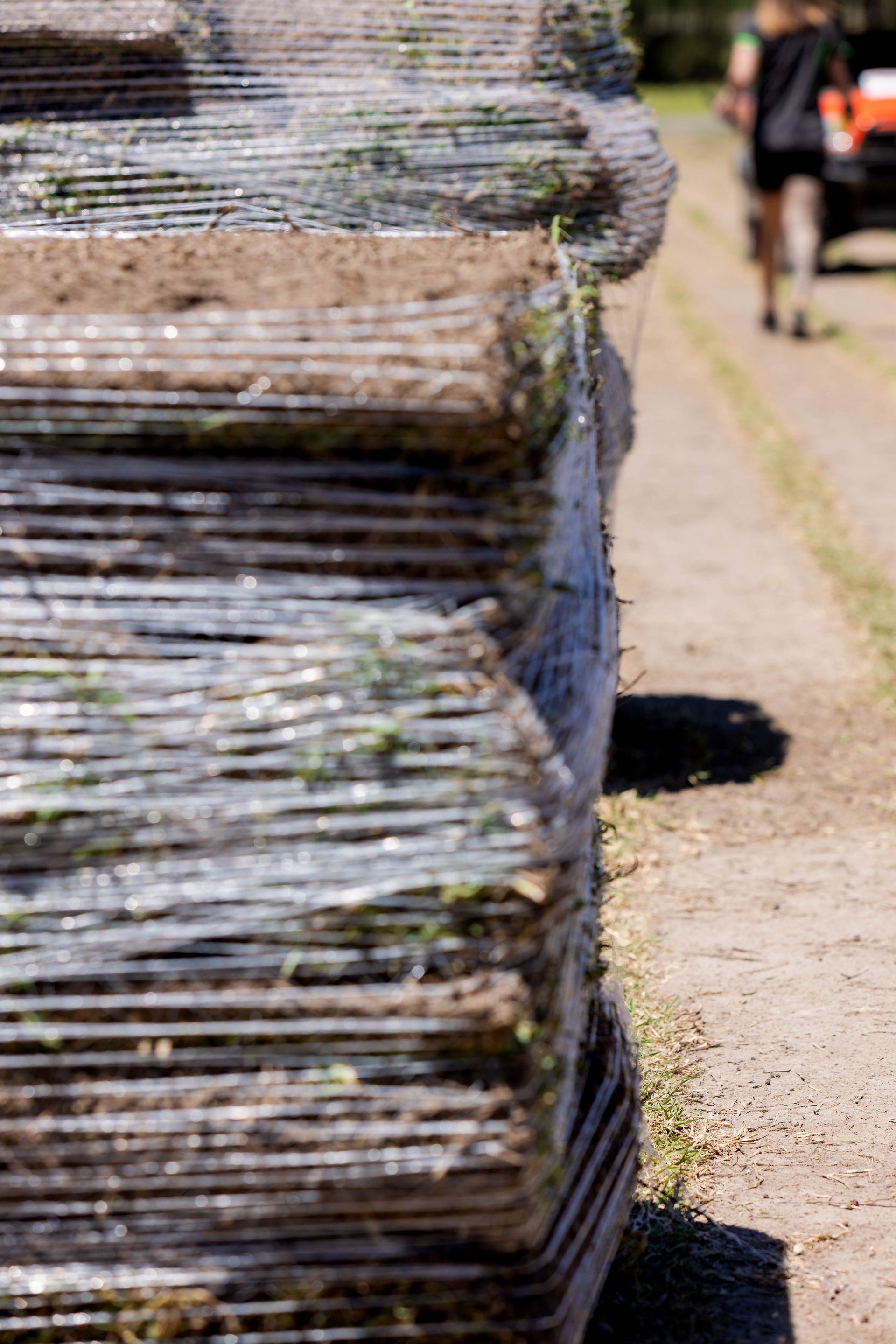 Stacked bundles of turf, wrapped in plastic, beside a dirt path with a person walking away.