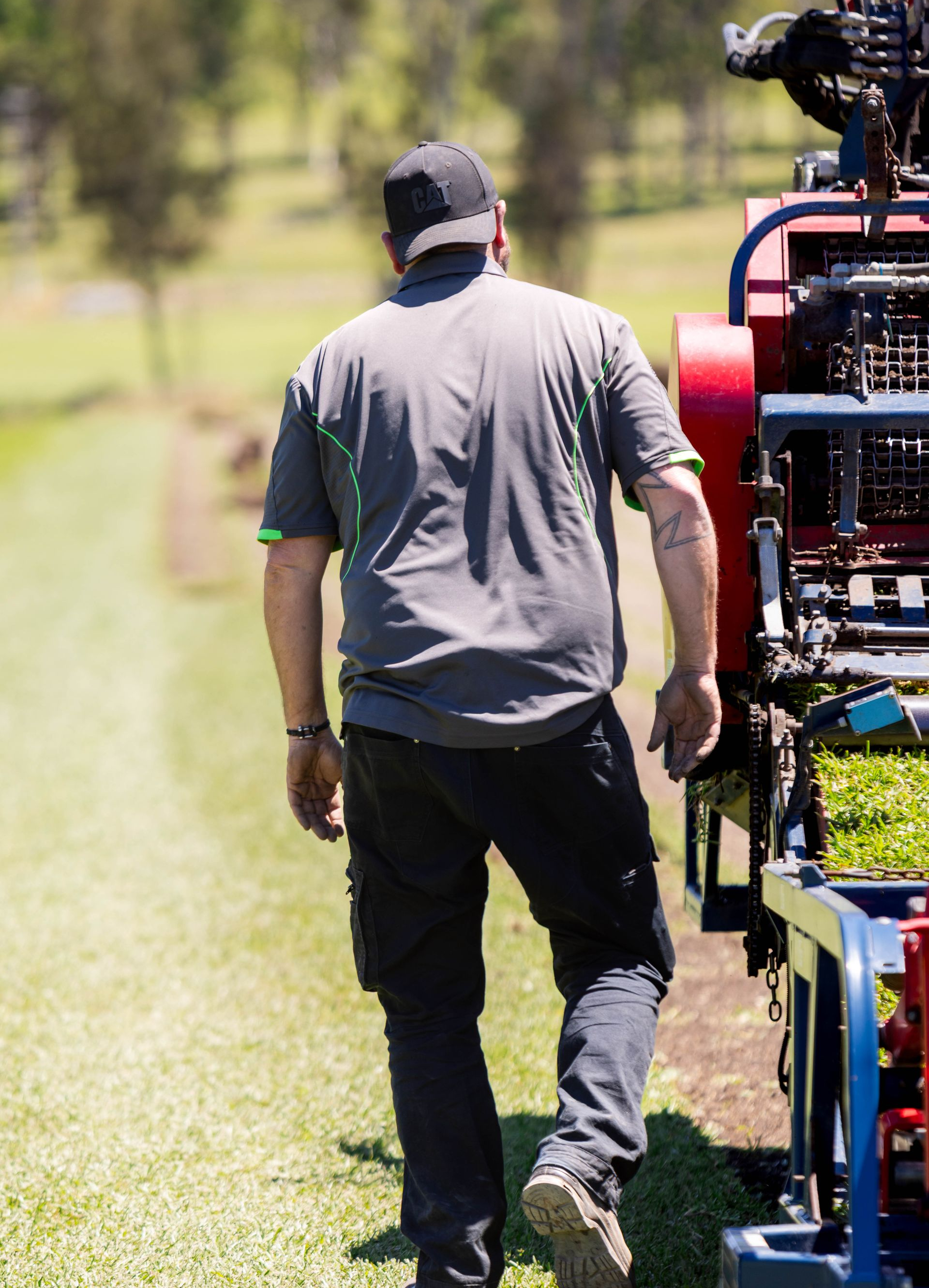 Man in gray shirt and black pants walks beside a machine laying sod on green grass.