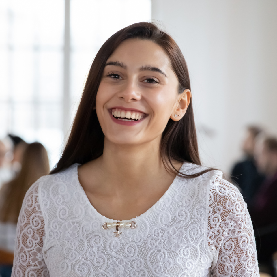 Young woman with long dark hair, wearing a white lace top, smiles widely