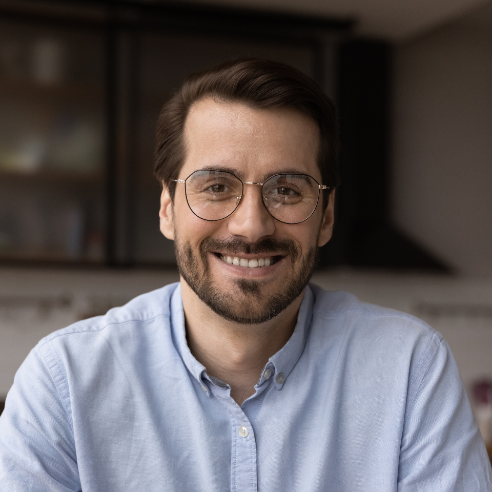 Man with glasses and a beard smiles broadly, wearing a blue button-down shirt. Indoor setting.