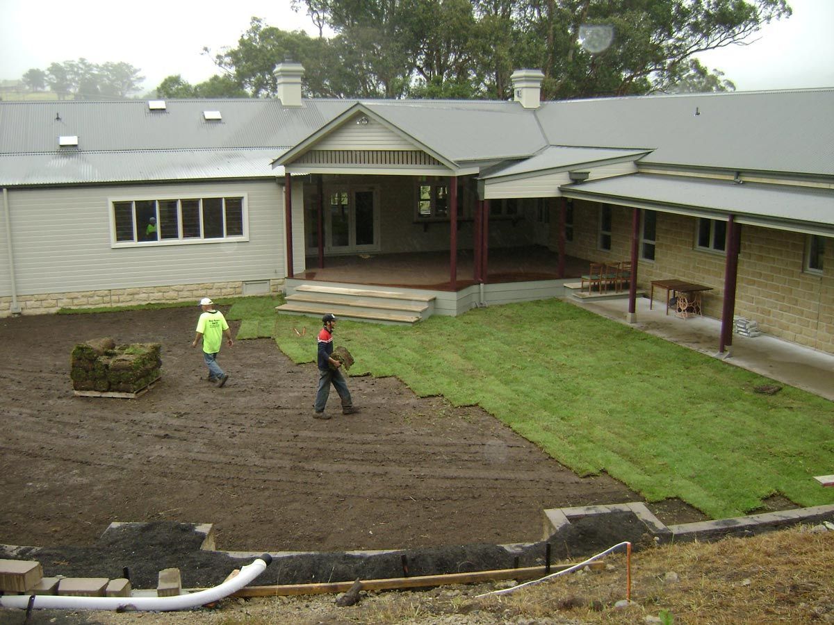 Two workers laying turf in a yard next to a building. One wears a safety vest, and the yard has a dark soil base.