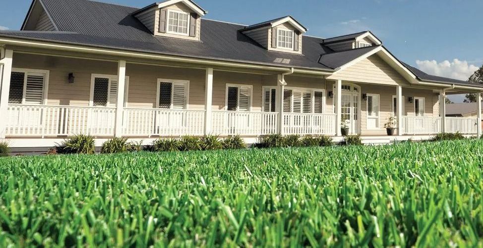A beige house with a porch and white railing sits in front of a vibrant green lawn under a blue sky.