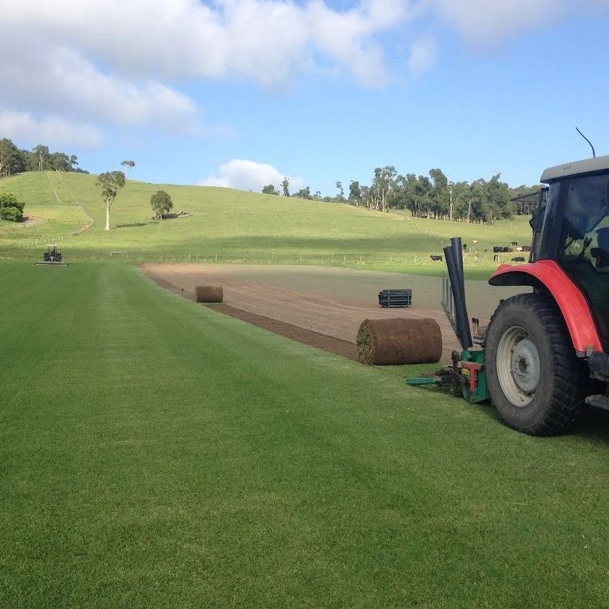 A tractor laying sod on a grassy field, with a partially laid strip and rolling hills in the background under a blue sky.