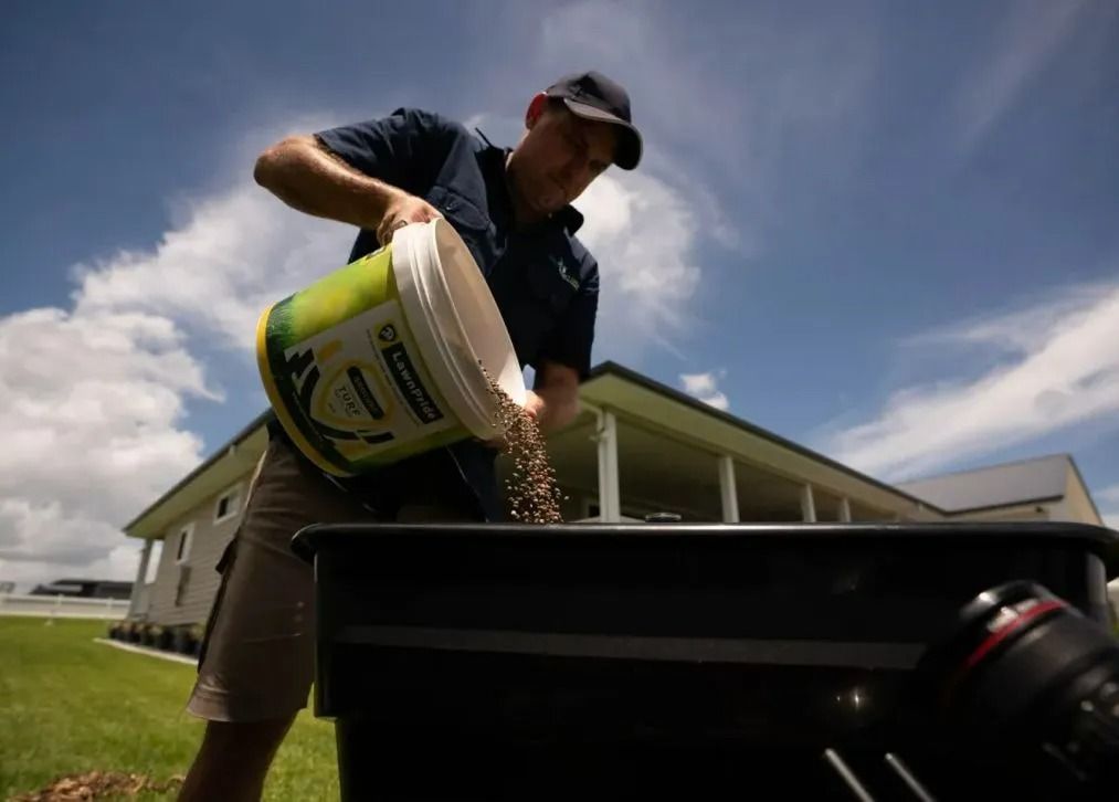 A man in a cap and shorts pours granules from a bucket into a black container, outside a house on a sunny day.